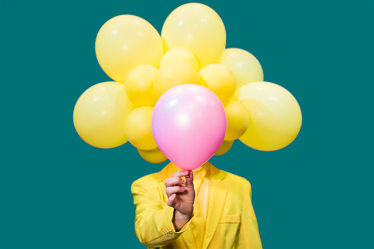 Keltaiseen pukeutunut ilmapallopäinen hahmo peittää kasvonsa vaaleanpunaisella ilmapallolla