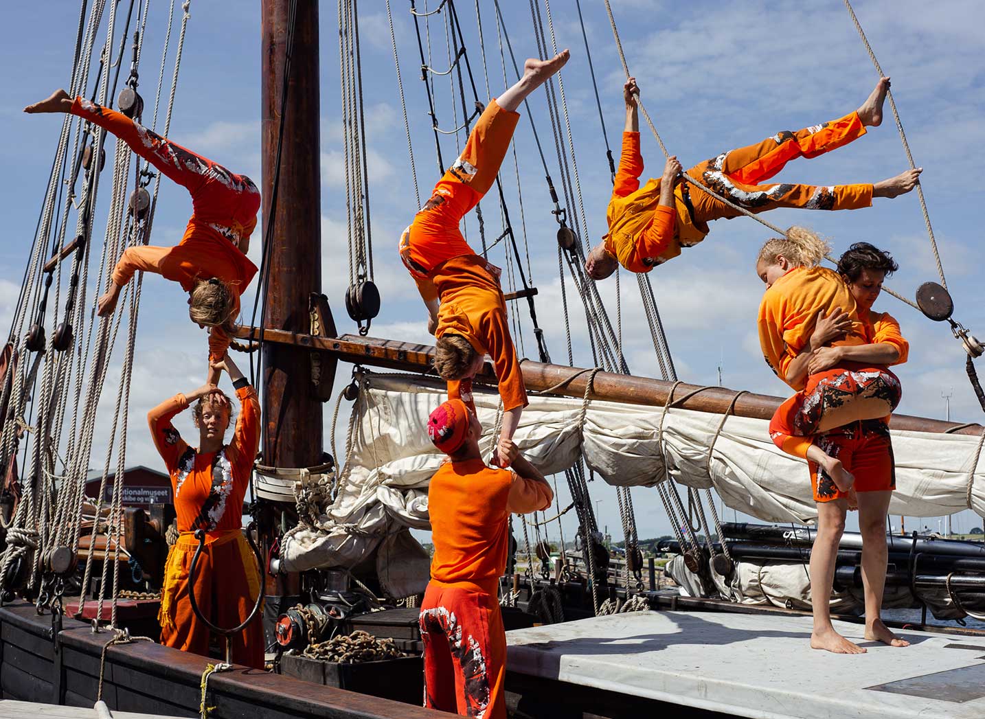 Seitsemän sirkustaiteilijaa laivan kannella, osa käsillään toisten taiteilijoiden käsien varassa.