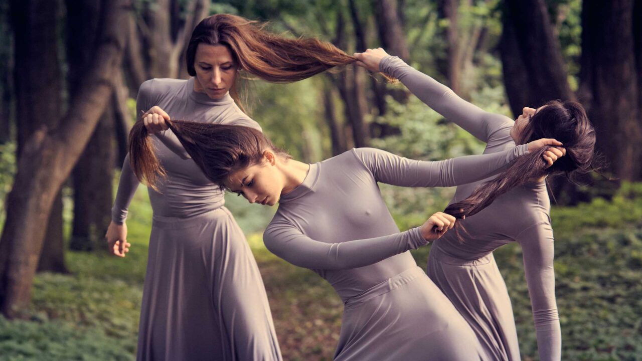 Kolme naista metsässä pitkissä harmaissa mekoissa pitelee toisiaan pitkistä hiuksista kiinni.
