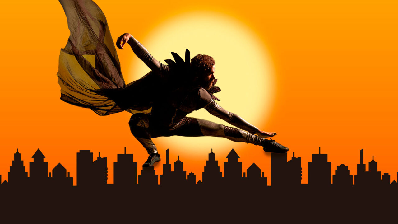 Supersankarin tumma hahmo kaupungin siluetin yllä, taustalla auringonlasku.